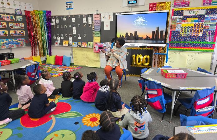  Students sitting on the floor in a classroom looking at their teacher holding reading a book