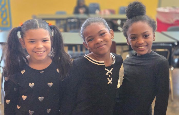 Three female students and all black, smiling and hugging one another.