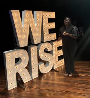 Attendance Agent Ms. Mason stands next to the we rise sign holding her nominee award.
